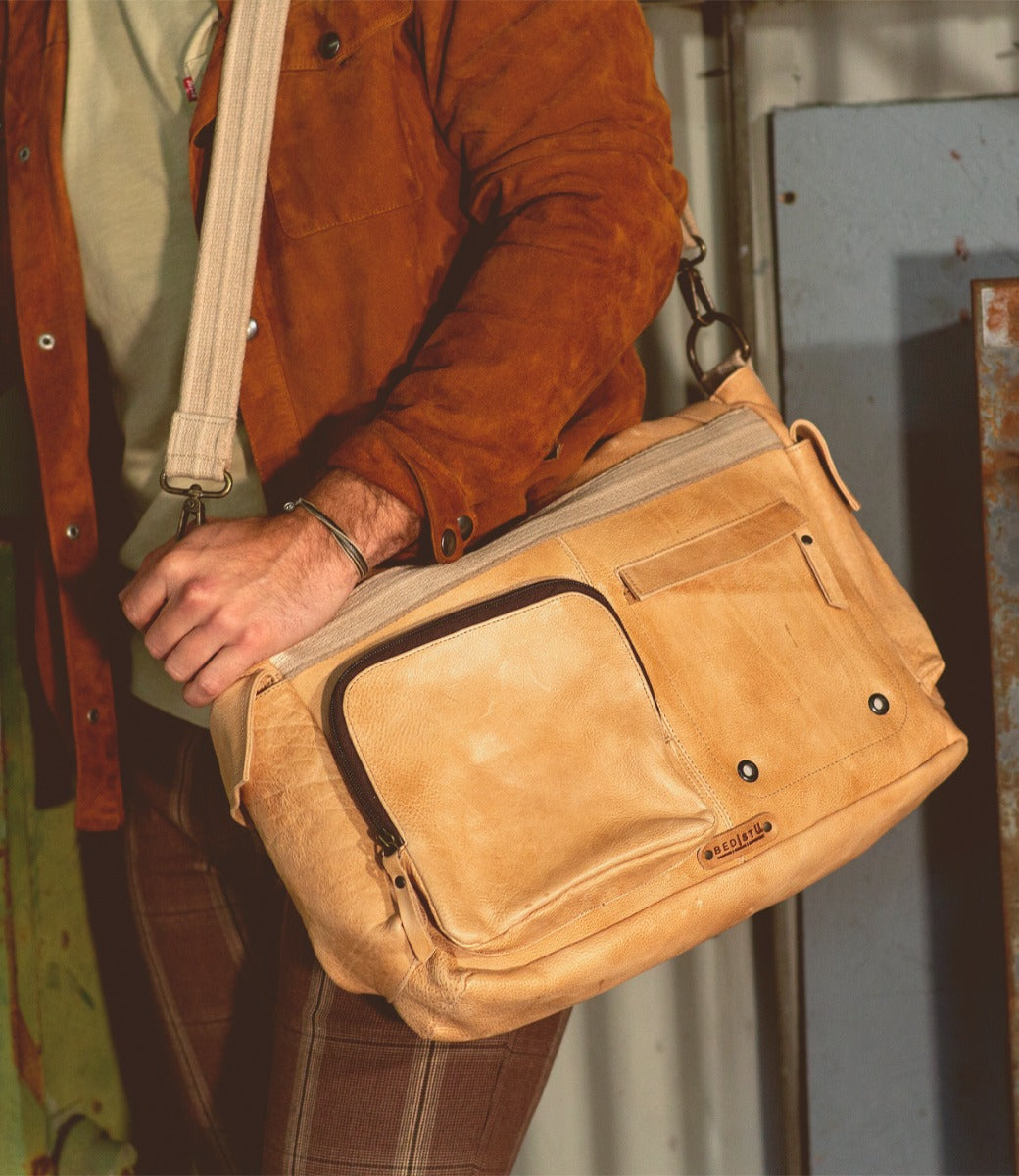A man is holding a Bed Stu Hawkeye II tan leather messenger bag.