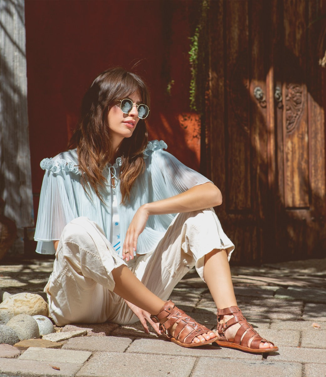 A woman is sitting on the ground in a pair of Bed Stu Hera sandals.