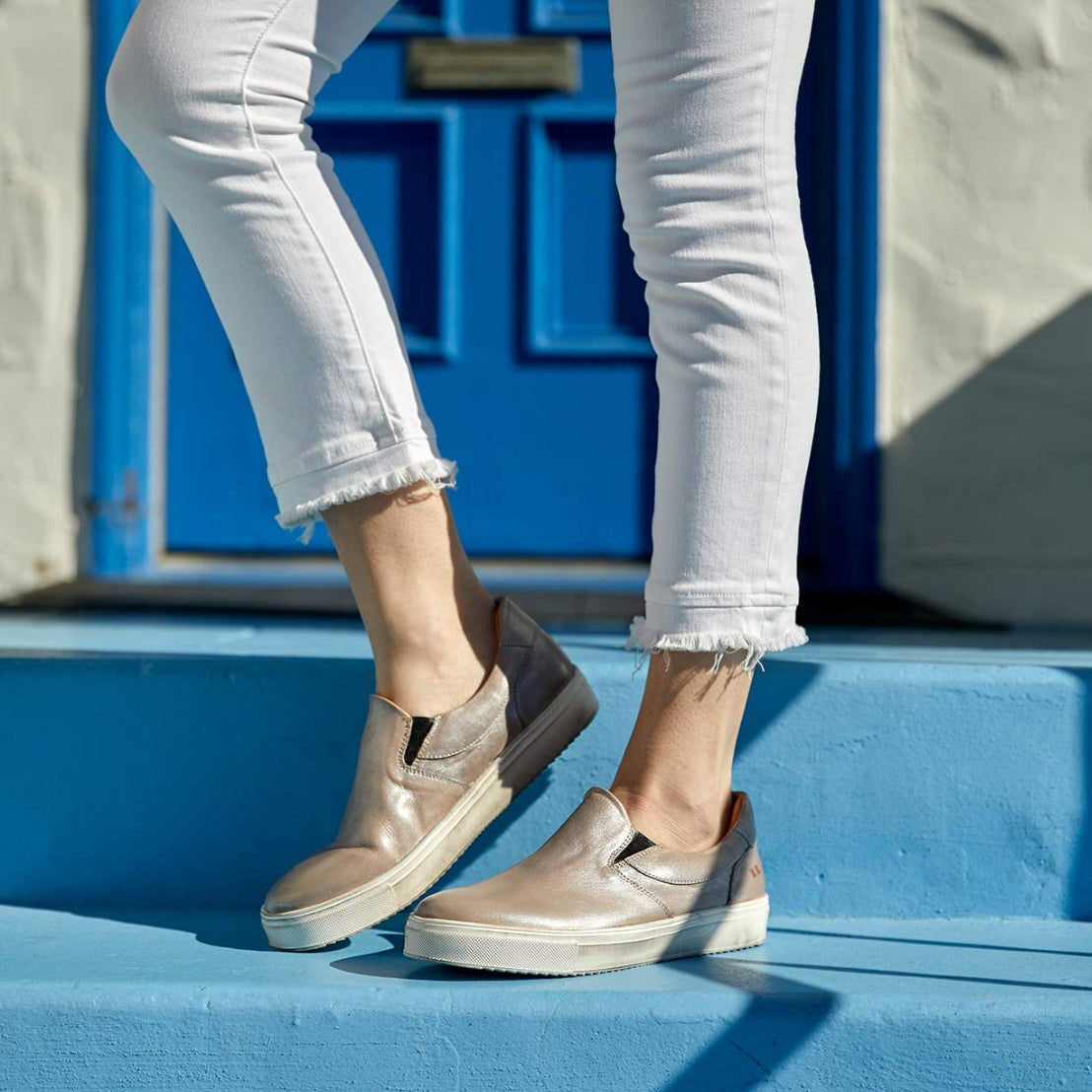 A woman wearing white jeans and tan Bed Stu slip on sneakers standing on a blue door.