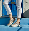A woman wearing white jeans and tan Bed Stu slip on sneakers standing on a blue door.