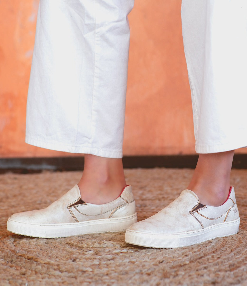 A woman wearing white pants and white Bed Stu Hermione slip on sneakers.
