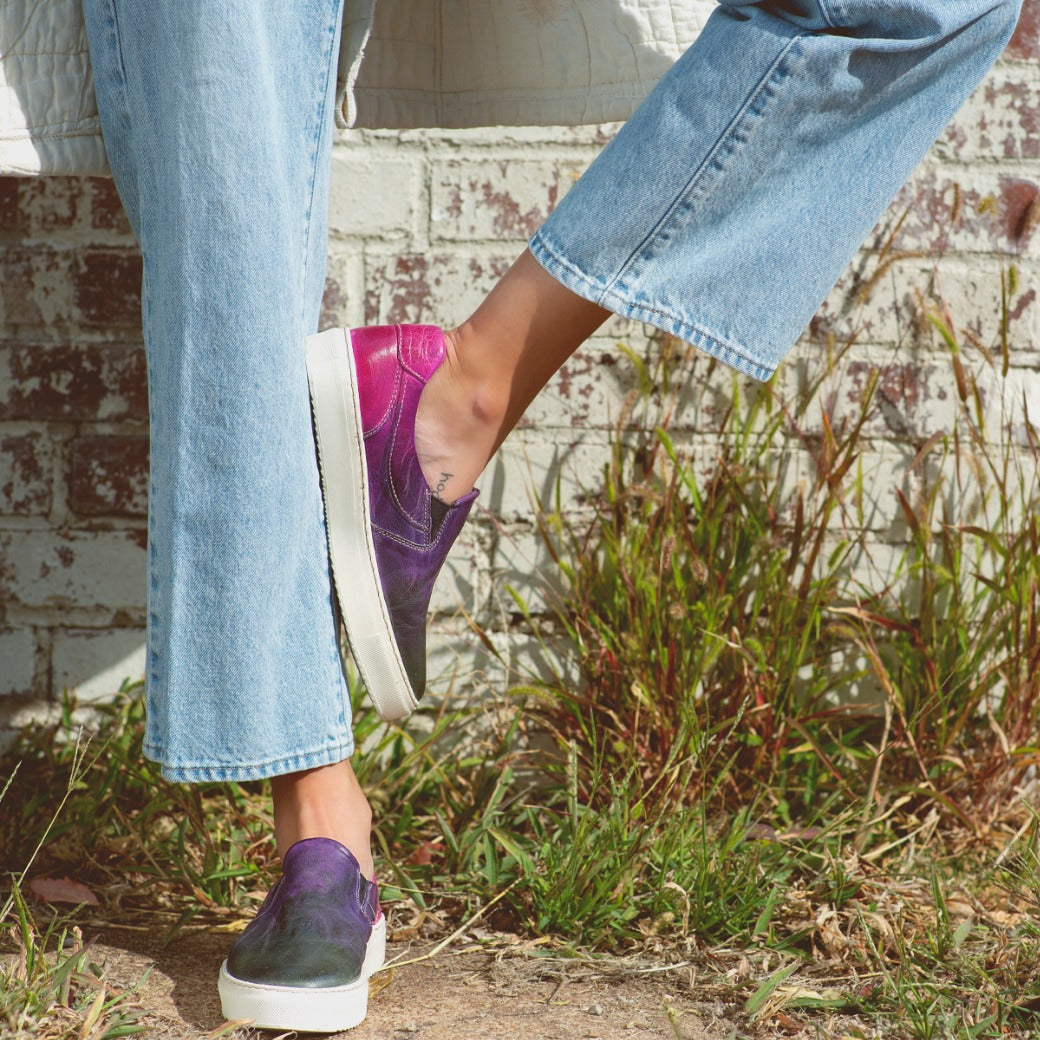 A woman wearing Hermione jeans and Bed Stu sneakers leaning against a brick wall.