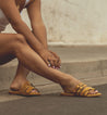 A woman sitting on a sidewalk wearing yellow Bed Stu Hilda sandals.