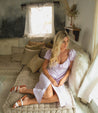 A woman in a lavender dress sitting on a couch wearing Hilda sandals from Bed Stu.