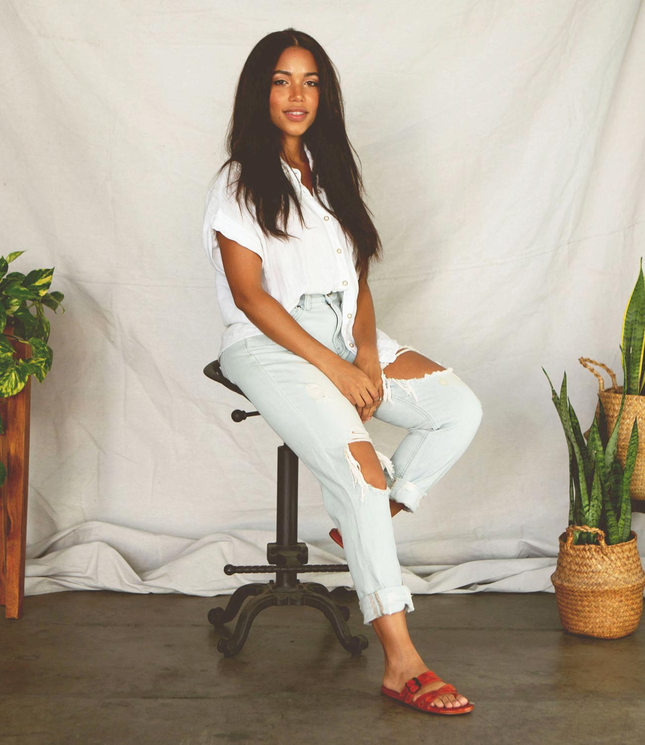 A woman sitting on a stool wearing ripped jeans, white shirt and a Hilda sandals from Bed Stu.