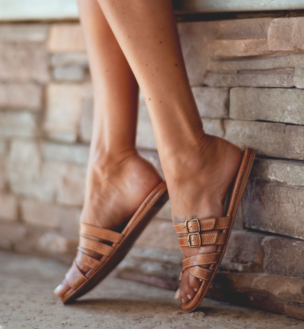 A woman wearing Hilda leather sandals by Bed Stu on a stone wall.