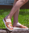 A woman wearing Bed Stu tan Holland sandals on a wooden bench.