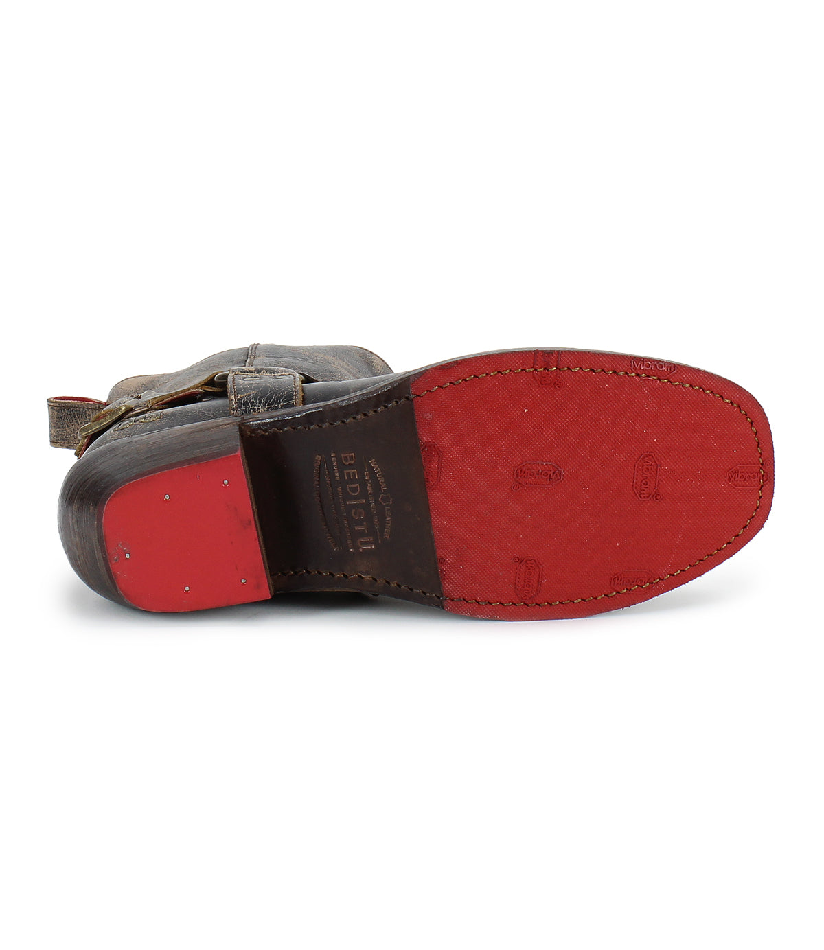 The image displays the bottom view of the Bed|Stü Homage boot, featuring brown leather construction with a red sole, visible stitching, and metal nails near the heel. The unique square toe design enhances its classic harness strap detailing with a stylish flair.