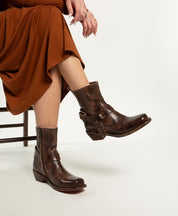 A person in a rust-colored dress sits on a chair, highlighting Bed Stu's Homage mid-calf brown leather harness boots with buckles and zippers against a white background.