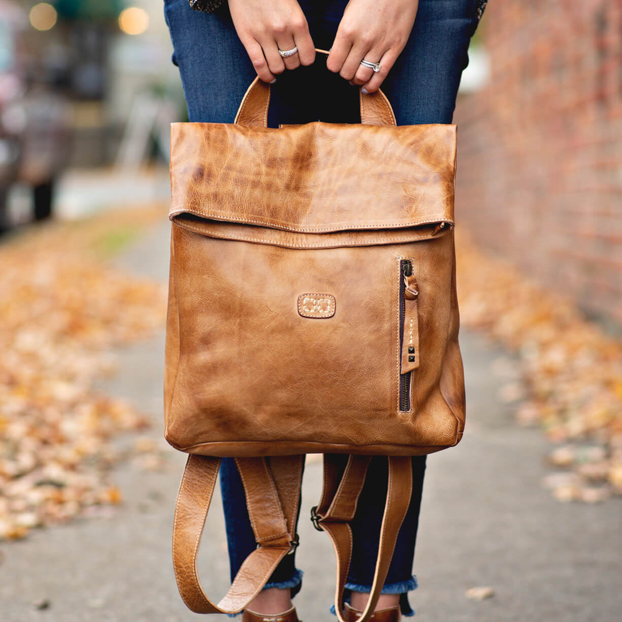 A woman holding a tan leather Bed Stu Howie backpack on a sidewalk.