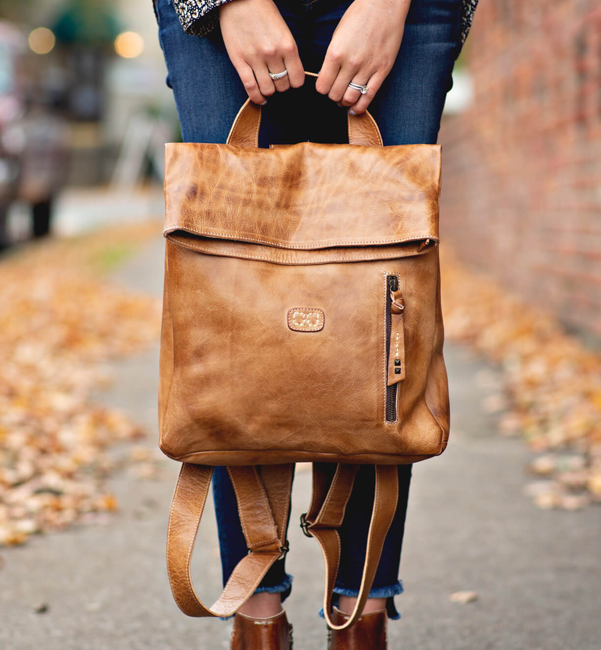 A woman holding a tan leather Bed Stu Howie backpack on a sidewalk.