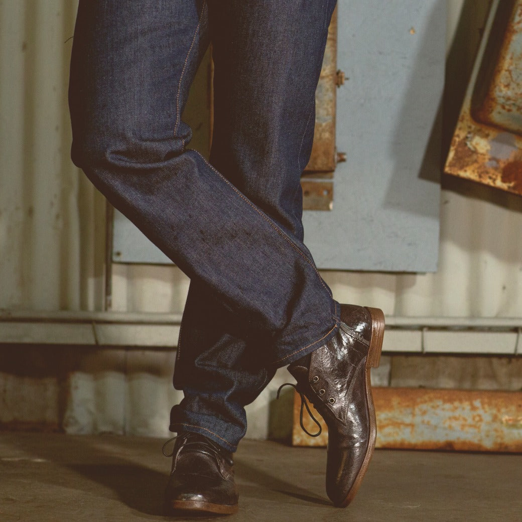 Close-up of a person's lower legs and feet, wearing dark jeans and brown leather Bed Stu Illiad chukka boots, standing in a dimly lit, industrial setting.