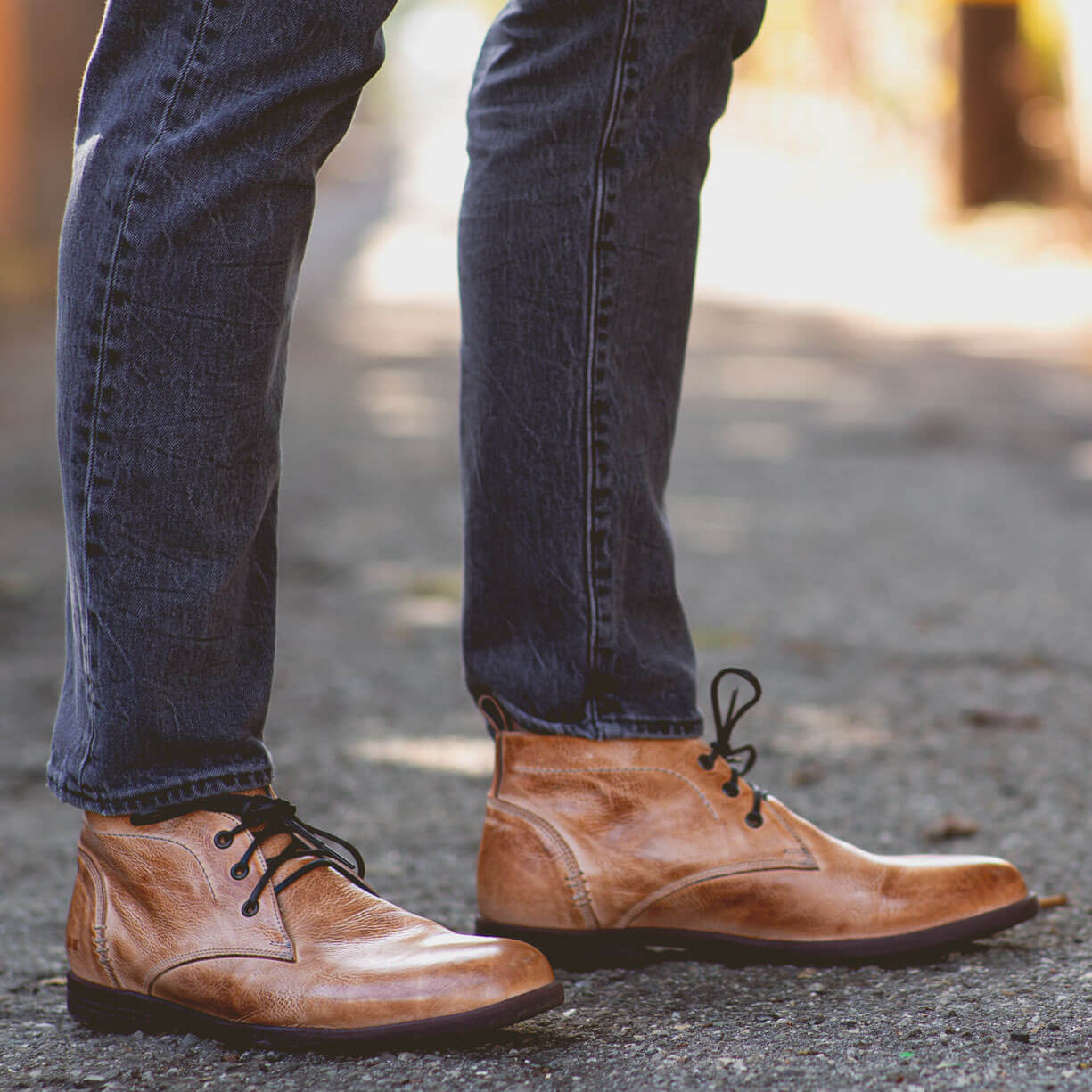 Close-up of a person's lower legs, wearing denim jeans and brown leather Bed Stu Illiad boots, standing on a gravel path.