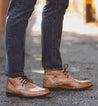 Close-up of a person's lower legs, wearing denim jeans and brown leather Bed Stu Illiad boots, standing on a gravel path.