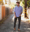 A man with a beard and tattoos wearing a striped shirt and jeans stands thoughtfully in a sunlit alley, his feet clad in Bed Stu Illiad boots.