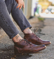 A person sitting on a curb, wearing Bed Stu Illiad boots and gray jeans, with a visible ankle tattoo that reads "free.