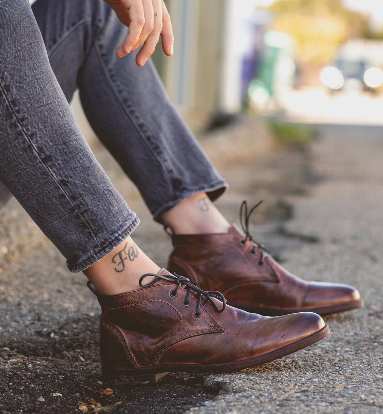 A person wearing Bed Stu Illiad Teak Rustic Boots and gray jeans sitting with one foot on the ground and one bent at the knee, showcasing a tattoo on their ankle.