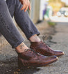 A person sitting on a curb, wearing Bed Stu Illiad boots and gray jeans, with a visible ankle tattoo that reads "free.