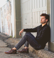 A man sitting on the ground, leaning against a wall, wearing a black shirt, jeans, and Bed Stu Illiad leather chukka boots made of vegetable-tanned leather, looking thoughtfully to the side.