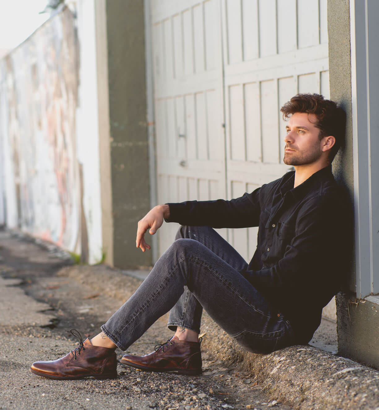 A man sitting on the ground, leaning against a wall, wearing a black shirt, jeans, and Bed Stu Illiad leather chukka boots made of vegetable-tanned leather, looking thoughtfully to the side.