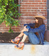 A woman wearing the Bed Stu Imelda cowboy hat and jeans sitting on a wall.