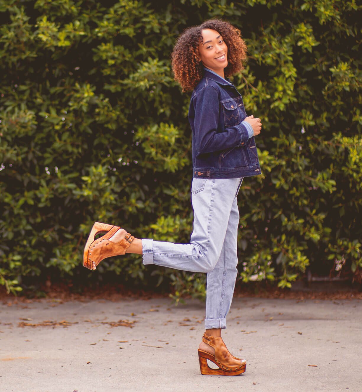 A woman in Bed Stu jeans and a Bed Stu denim jacket posing in front of bushes.