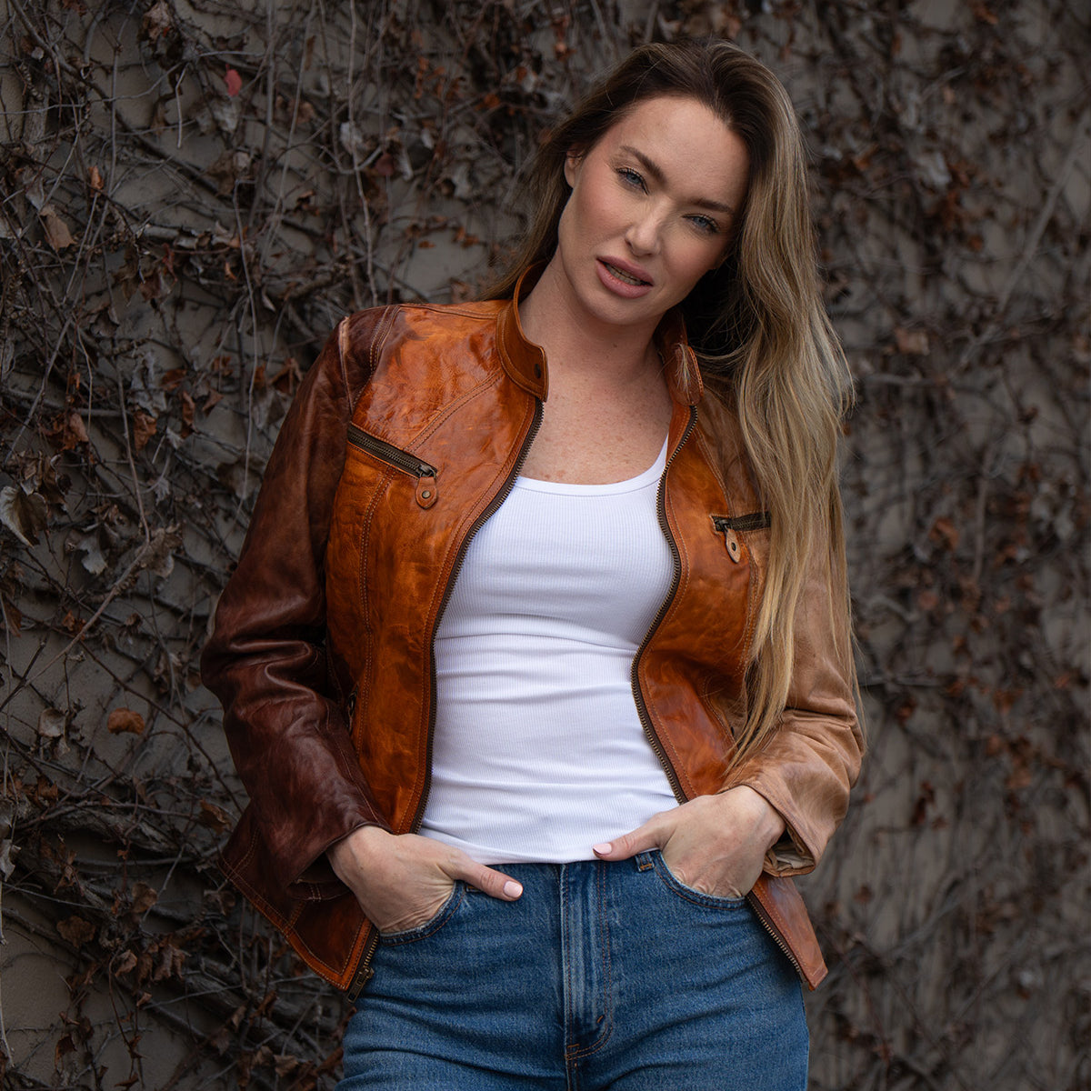 A woman in a Bed Stu Include handcrafted leather jacket and blue jeans stands against a wall of bare, tangled vines, looking at the camera.