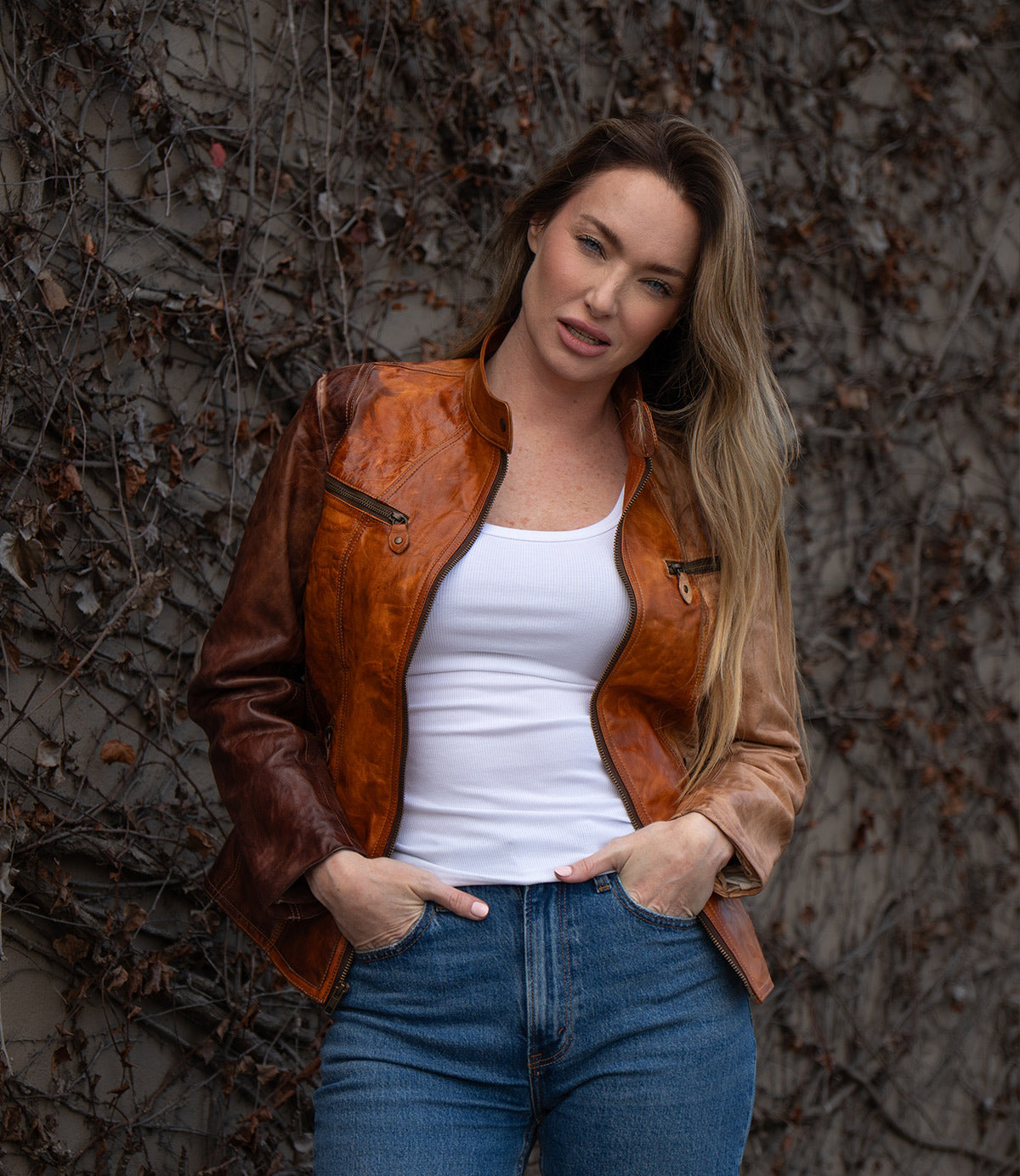 A woman in a Bed Stu Include handcrafted leather jacket and blue jeans stands against a wall of bare, tangled vines, looking at the camera.