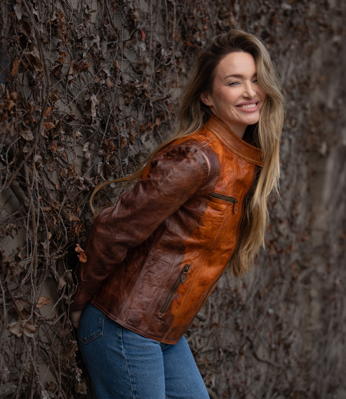 A woman wearing the Bed Stu Include handcrafted leather jacket and blue jeans stands smiling in front of a wall covered with dry vines, facing the camera.