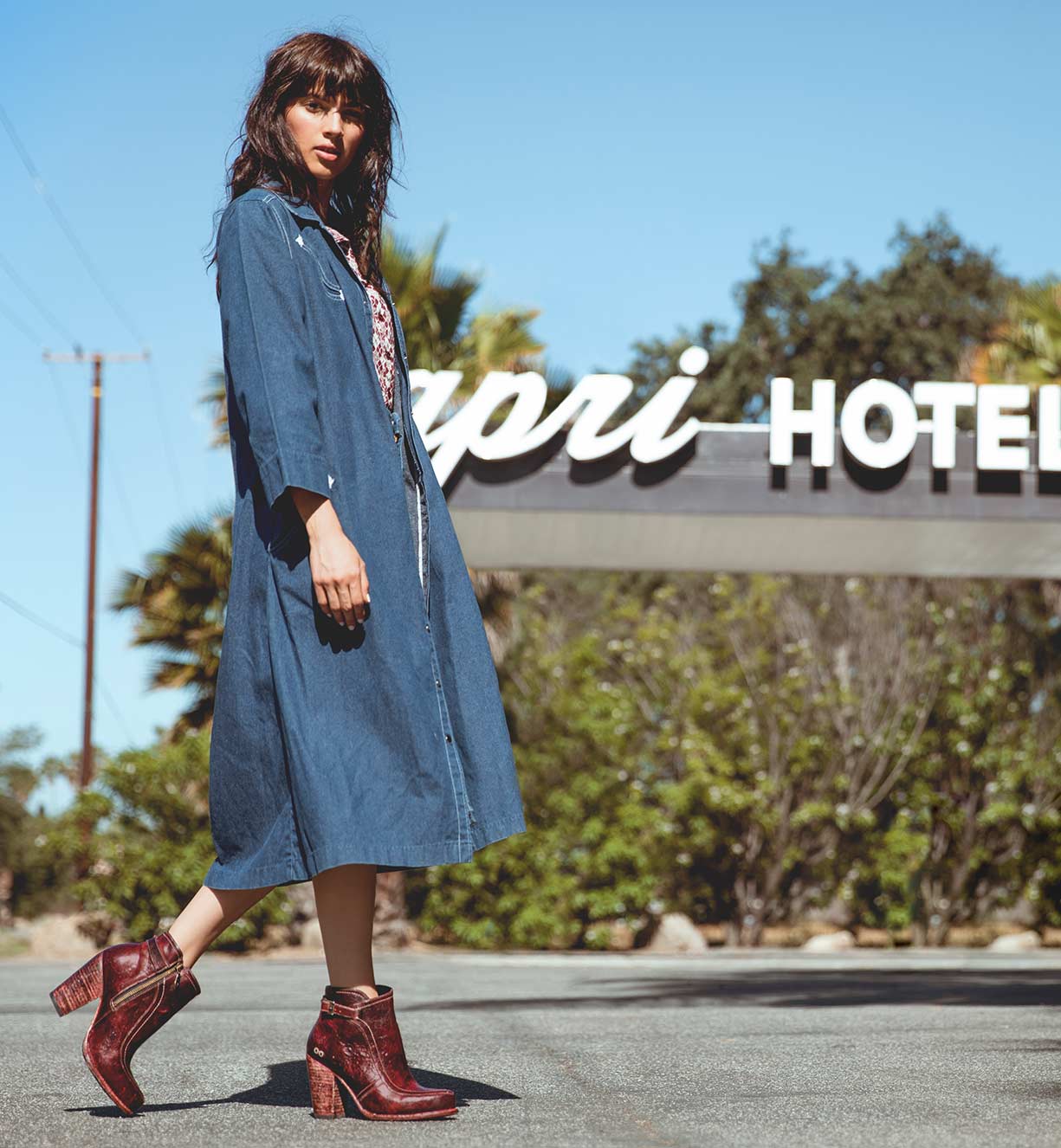 A woman in an Isla denim dress standing in front of a hotel sign. (Brand: Bed Stu)
