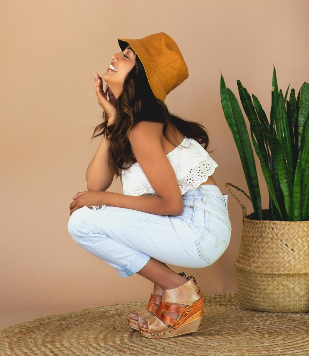 A woman wearing a Jacey II wedge sandals, tan hat and jeans crouching in front of a potted plant.