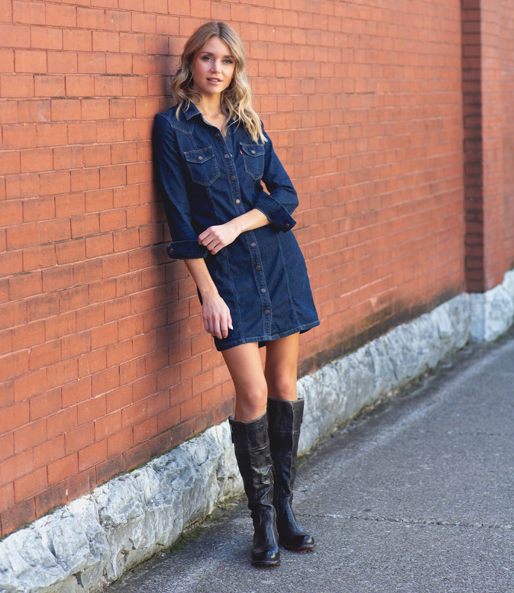 A woman in a denim shirt dress leaning against a brick wall wearing the Jacqueline by Bed Stu.