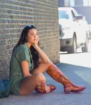A woman is sitting on the sidewalk in a green dress and knee high Bed Stu Jacqueline boots.