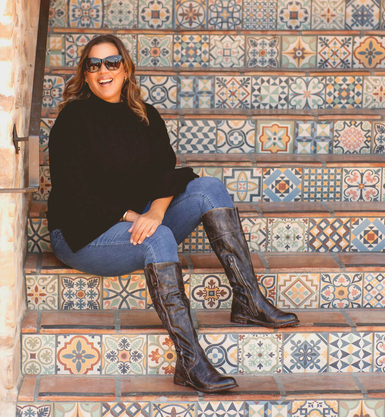 A woman wearing a pair of Jacqueline Wide Calf boots by Bed Stu and a black sweater sits on a set of stairs.