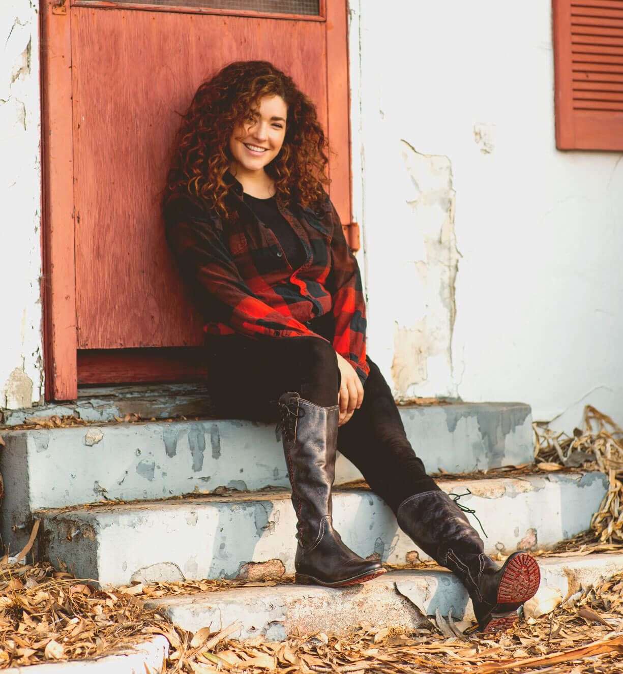 A woman in a plaid shirt sits on the steps of an old house wearing Janina shoes by Bed Stu.