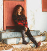 A woman in a plaid shirt sits on the steps of an old house wearing Janina shoes by Bed Stu.