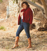 A woman in a red Janina jacket and jeans posing in front of a tree, carrying a Bed Stu bag.