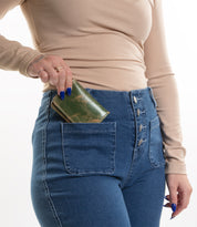 Wearing a beige long-sleeve shirt and high-waisted blue jeans, a person slips the Bed Stu Jeor compact wallet, featuring handy credit card slots, into their front pocket.