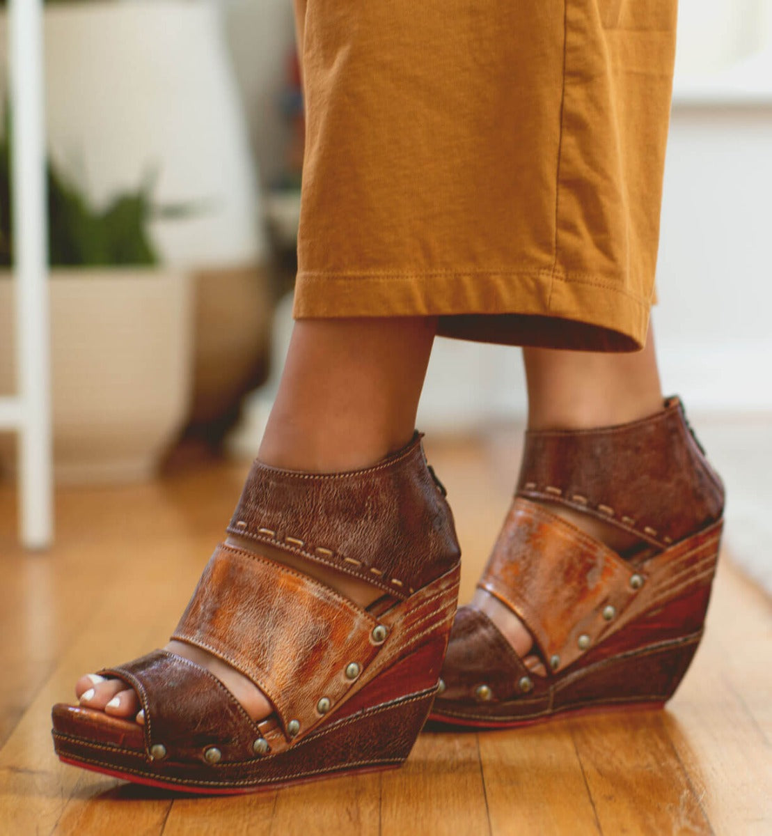 A woman's feet standing on a wooden floor in Bed Stu Jessie brown wedge sandals.
