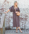 A blonde woman in a striped dress, leaning against a brick wall while carrying a Bed Stu Joshebed crossbody handbag.