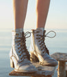 A woman's legs standing on a railing next to the ocean, wearing the Judgement shoes by Bed Stu.