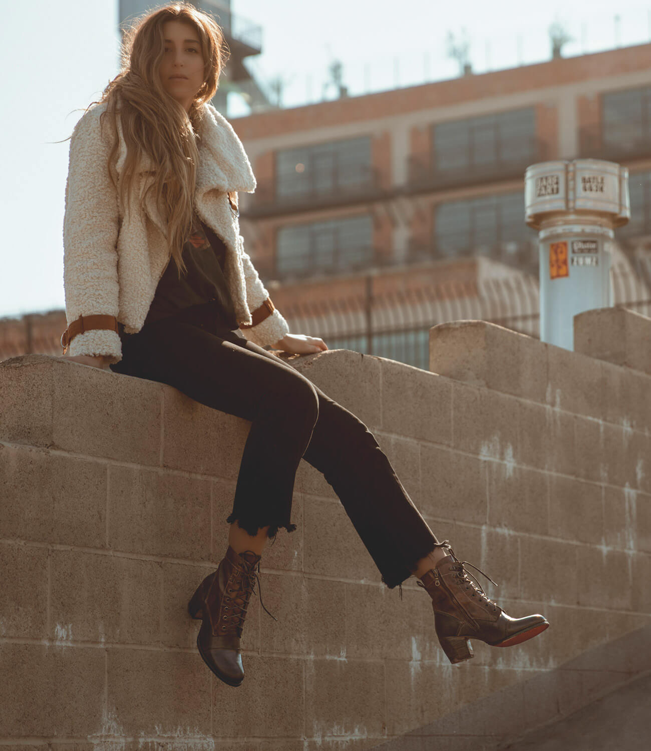 A woman is sitting on a wall wearing a Bed Stu Judgement jacket and boots.