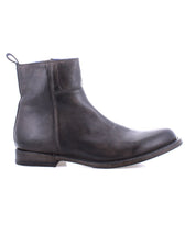 The Bed Stu Kaldi is a black leather ankle boot featuring a side zipper, mid-height heel, and a convenient pull tab at the back. The boot boasts a slightly worn look with visible stitching, highlighting its handmade quality and classic silhouette.