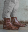 A person wearing cuffed gray jeans and Bed Stu's Kaldi brown leather ankle boots with zippers, standing on a concrete floor.