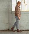 Man in a brown jacket and Bed Stu leather ankle boot, walking past a brick wall.