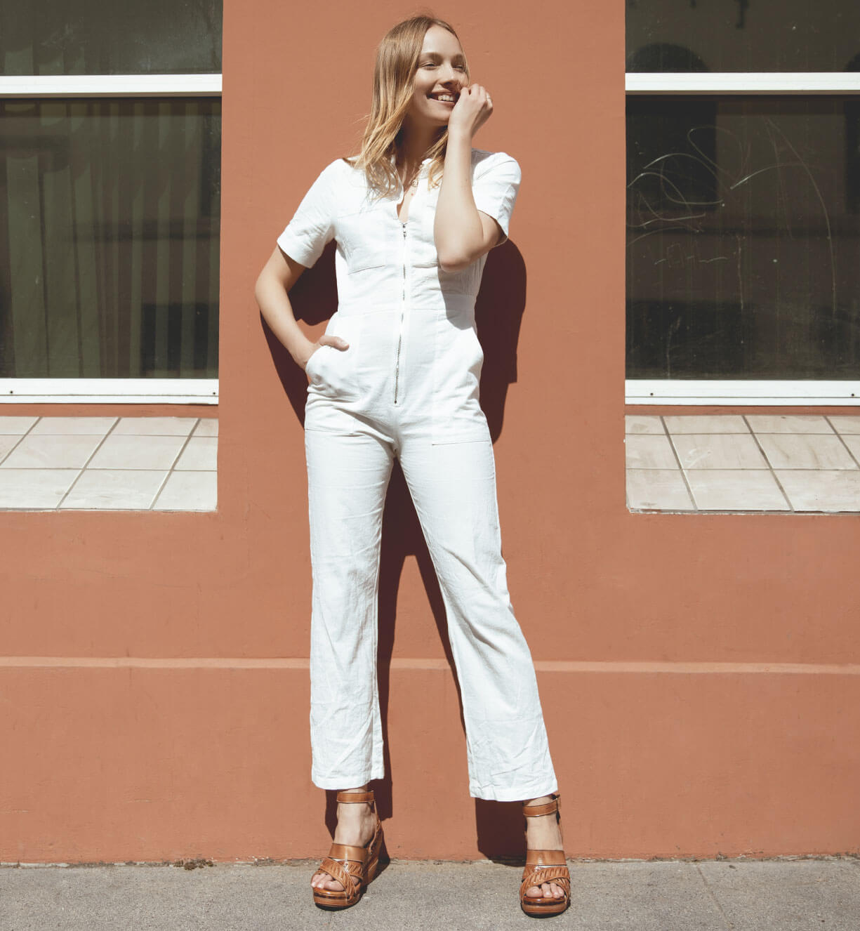 A woman in a white Kaphie jumpsuit leaning against a wall. Brand: Bed Stu