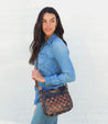 A woman wearing a denim shirt, jeans, and black and brown woven Keiki bag while standing next to a white wall.