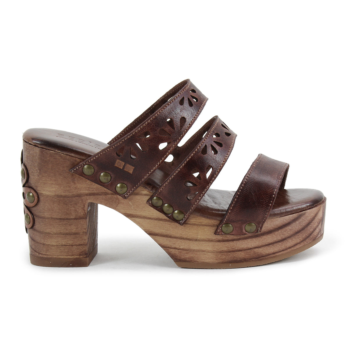 The Kreep by Bed Stu is a brown platform sandal featuring a natural wooden block heel, three leather straps with floral cutouts, and brass studs, evoking a 70’s-inspired look.