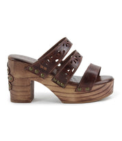 The Kreep by Bed Stu is a brown platform sandal featuring a natural wooden block heel, three leather straps with floral cutouts, and brass studs, evoking a 70’s-inspired look.