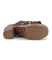 The Kreep by Bed Stu is a 70's-inspired sandal with brown leather straps and natural wooden heels. It features intricate nail and rivet details, while delicate floral cutouts seen from the side enhance its timeless design.